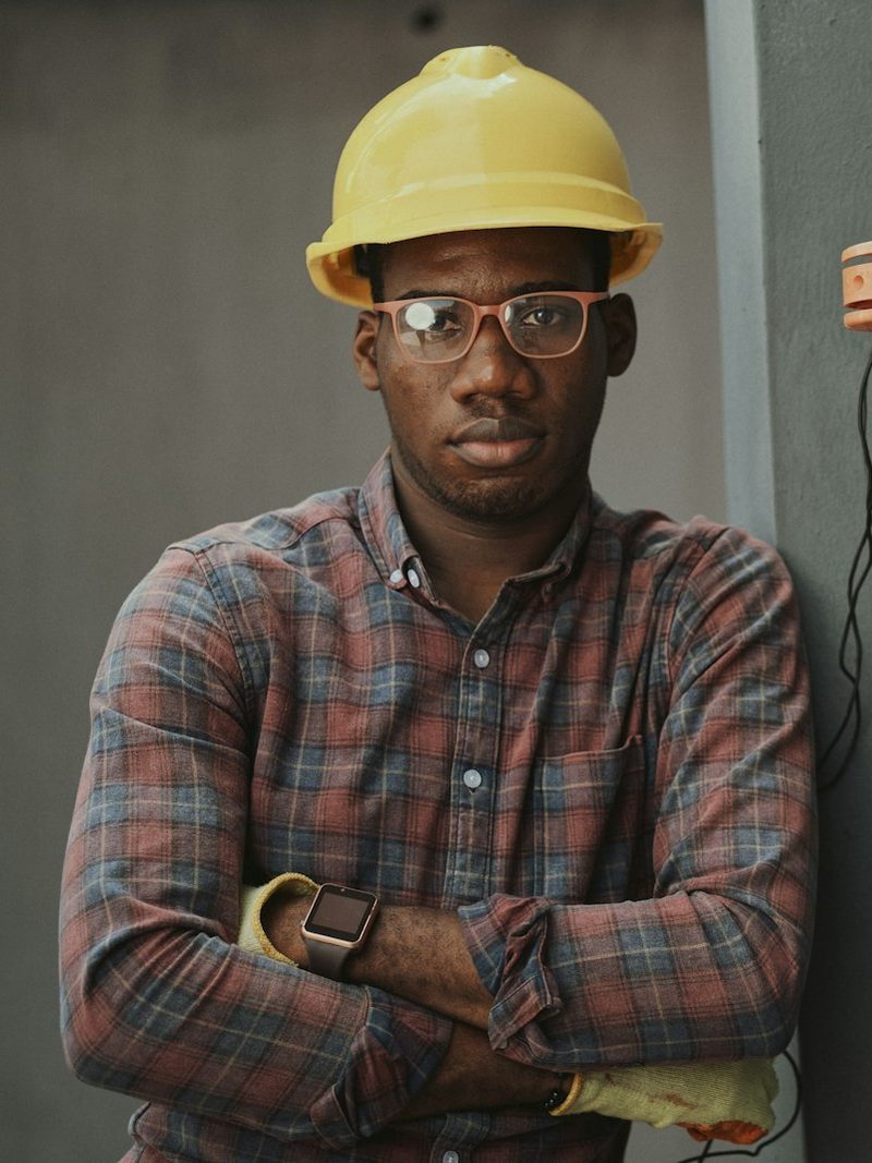 Portrait von Ali, Geselle, mit gelbem Schutzhelm, Brille und kariertem Hemd neben einem Stromkasten